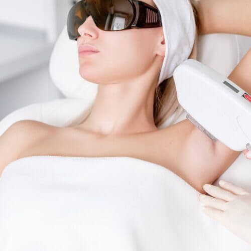 Young pretty caucasian woman in dark glasses is undergoing photoepilation armpits procedure in beauty clinic. Concept removal of unwanted hair from body skin. Intimate area hygiene